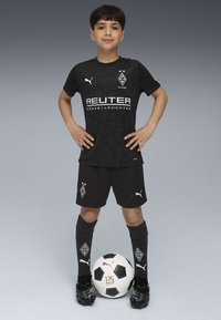 Junge in schwarzem Borussia Mönchengladbach Fußballtrikot steht mit auf die Hüften gestützten Händen und einem Fußball zu seinen Füßen vor grauem Hintergrund.