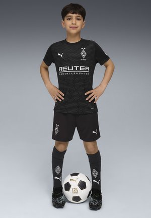 Chłopiec w czarnej koszulce piłkarskiej Borussia Mönchengladbach stoi z rękami na biodrach i piłką u stóp na szarym tle.