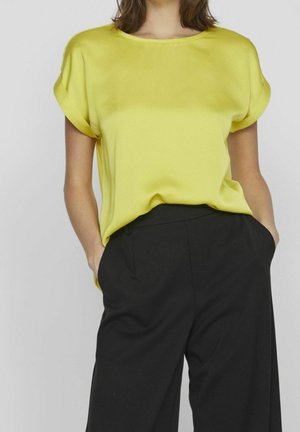 Keltainen satiininen lyhythihainen pusero, jossa on pyöreä pääntie, yhdistetty korkeavyötäröisiin mustiin housuihin. Silkkinen pinta ja rento istuvuus.