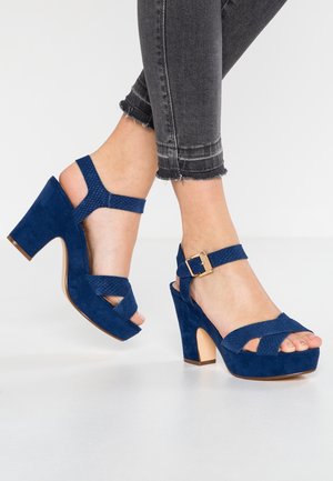 Pieds chaussés de sandales à talons hauts en daim bleu avec des brides à la cheville et des boucles dorées, assorties à un jean noir raccourci à ourlets effilochés.
