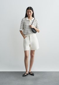 Πλεχτό πόλο μπλουζάκι με οριζόντιες ρίγες σε μαύρο και ά�σπρο, συνδυασμένο με κρεμ tailored βερμούδες. Μαύρη υφασμάτινη τσάντα και επίπεδα παπούτσια.