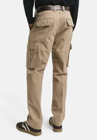 Beige Cargo-Hosen mit Seitentaschen, aus Baumwolle gefertigt, mit geradem Bein. Kombiniert mit dunklem Schuhwerk, von hinten betrachtet.