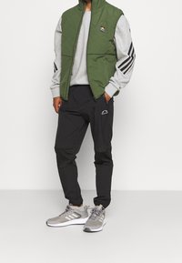 Gilet trapuntato verde sopra una felpa grigia, pantaloni neri con dettagli, e sneakers grigie con suole bianche. Presenta maniche a righe e loghi.