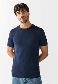 Vīrietis ar cirtainiem matiem un bārdu, valkājot tumši zilu vienkāršu īsroku t-kreklu un gaiši zilas džinsa bikses uz baltas sienas fona.