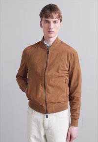Hnědá semišová bomber bunda s pružnými manžetami a límcem. Přední zip a boční kapsy. Nosená přes světle modrou košili a bílé kalhoty.
