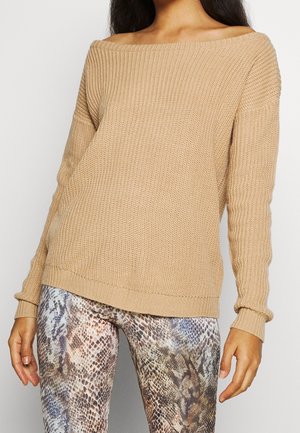 Kvinde iført en beige ribstrikket off-shoulder sweater kombineret med højttaljede bukser med slangeprint, stående mod en ensfarvet baggrund.