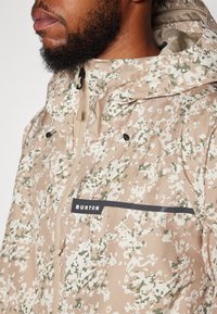 Veste à capuche avec un motif camouflage beige et vert, dotée d'un logo noir et de fermetures éclair. Texture lisse, matériau léger.