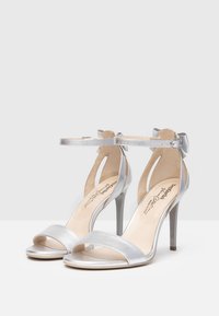 Sandales à talons hauts argentées avec une finition métallique, orteils ouverts, bride de cheville et détail en nœud à l'arrière. Texture lisse et design épuré.