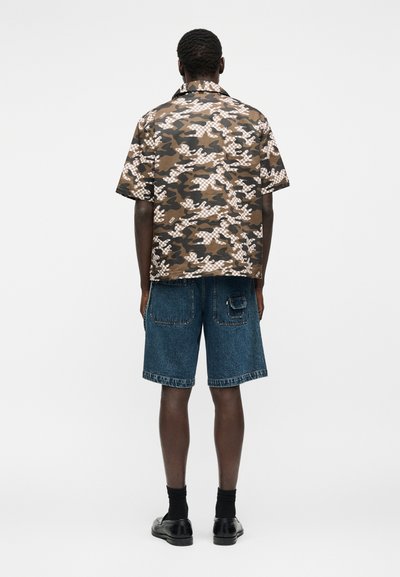 Άνδρας που στέκεται πλάτη, φοράει καφέ και μπεζ κοντομάνικο πουκάμισο καμουφλάζ, μπλε τζιν σορτς, μαύρες κάλτσες και μαύρα παπούτσια.