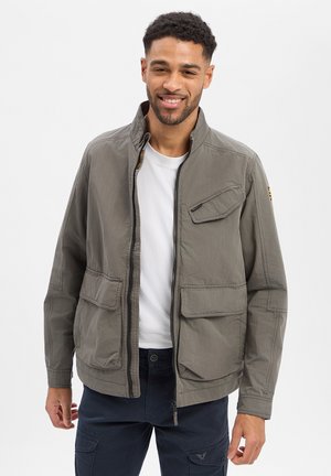 Man smiling, wearing gray zip-up jacket over white shirt and dark cargo pants, standing against plain light background.