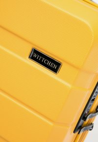 Valigia rigida gialla con superficie texturizzata, hardware nero e logo in rilievo. Presenta un design rigato per resistenza e stile.