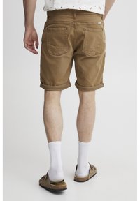 Braune Baumwollshorts mit umgeschlagenen Säumen, ausgestattet mit zwei Gesäßtaschen und dezenten Stickdetails. Getragen mit weißen Socken und Slipper-Sandalen.