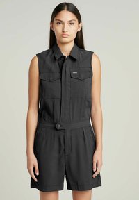 Tutina nera senza maniche con chiusura a bottoni, due tasche sul petto, vita con cintura e vestibilità comoda. Il tessuto sembra liscio con una texture minima.