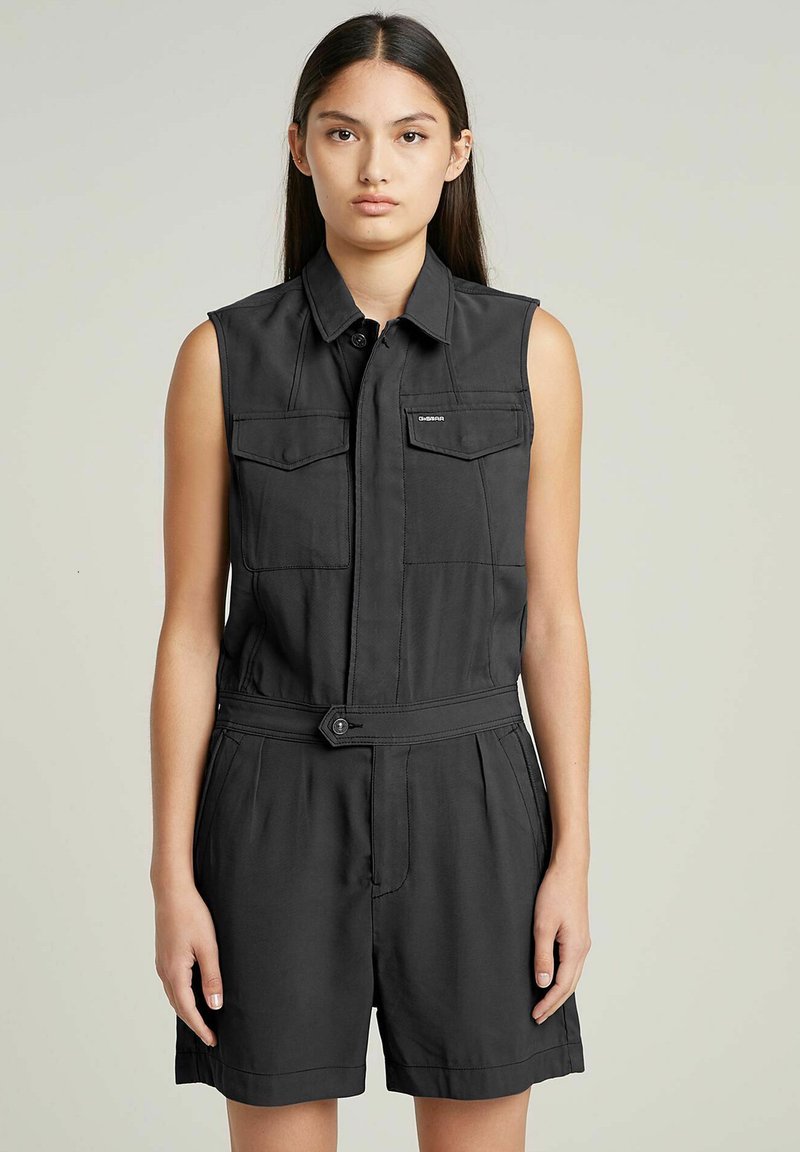 Tutina nera senza maniche con chiusura a bottoni, due tasche sul petto, vita con cintura e vestibilità comoda. Il tessuto sembra liscio con una texture minima.