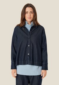 Mørk denimskjorte med knaplukning foran, lange ærmer og en afslappet pasform, lagd over en lyseblå ribstriktop.