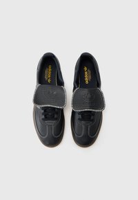 Sorte lærersneakers med hvite sømmer, rund tå og tykk gummisåle. Har en stor, teksturert svart tunge med merking.