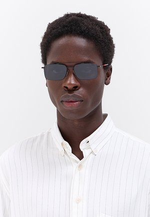 Giovane con capelli corti e ricci che indossa occhiali da sole rettangolari scuri e una camicia bianca abbottonata con sottili righe verticali.