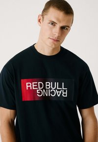 Melna kokvilnas T-krekls ar lielu sarkanu un baltu grafiku, kurā horizontāli un vertikāli ir drukāts "RED BULL RACING". Īsas piedurknes un apaļa kakla izgriezne.
