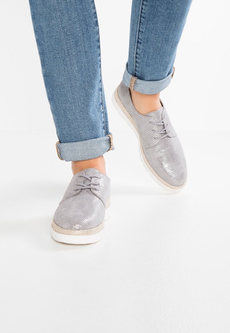 Chaussures grises à lacets en matériau texturé avec une finition brillante, dotées d'une semelle blanche et d'un accent en fibre naturelle autour de la semelle.