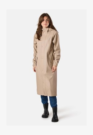 Vrouw draagt een beige lange regenjas met capuchon en drukknopen, blauwe spijkerbroek en zwarte rubberlaarzen, staand op een witte achtergrond.