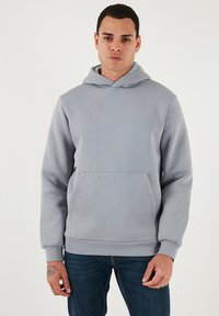 Világosszürke kapucnis pulóver, puha anyagból, első zsebbel, bordázott mandzsettával és húzózsinórral ellátott kapucnival. Sötétkék farmernadrággal viselve.