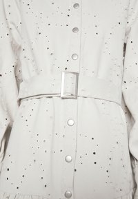 Robe en denim blanc avec ceinture à boucle rectangulaire argentée, œillets décoratifs et boutons-pression sur le devant.