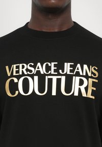 Črna dukserica z velikim zlatim napisom "VERSACE JEANS COUTURE" v izrazitem tisku. Gladka tekstura tkanine, dizajn s krojem okoli vratu.