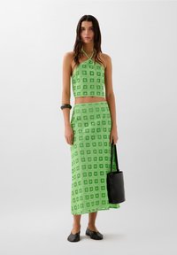 Zelená dvoudílná souprava skládající se z halter crop topu a dlouhé sukně s čtverečkovaným vzorem. Modelka drží černou kbelíkovou tašku a na nohách má černé baleríny.