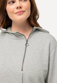 Felpa grigia con zip e cappuccio grande, caratterizzata da una cerniera nera, texture morbida e vestibilità ampia. Nessun motivo o accentuazione visibile.