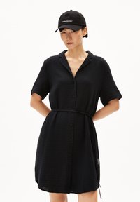 Schwarzes Hemdkleid mit kurzen Ärmeln, strukturiertem Stoff, Gürtel in der Taille und einem Kragen. Ergänzt durch eine schwarze Mütze mit weißem Text.