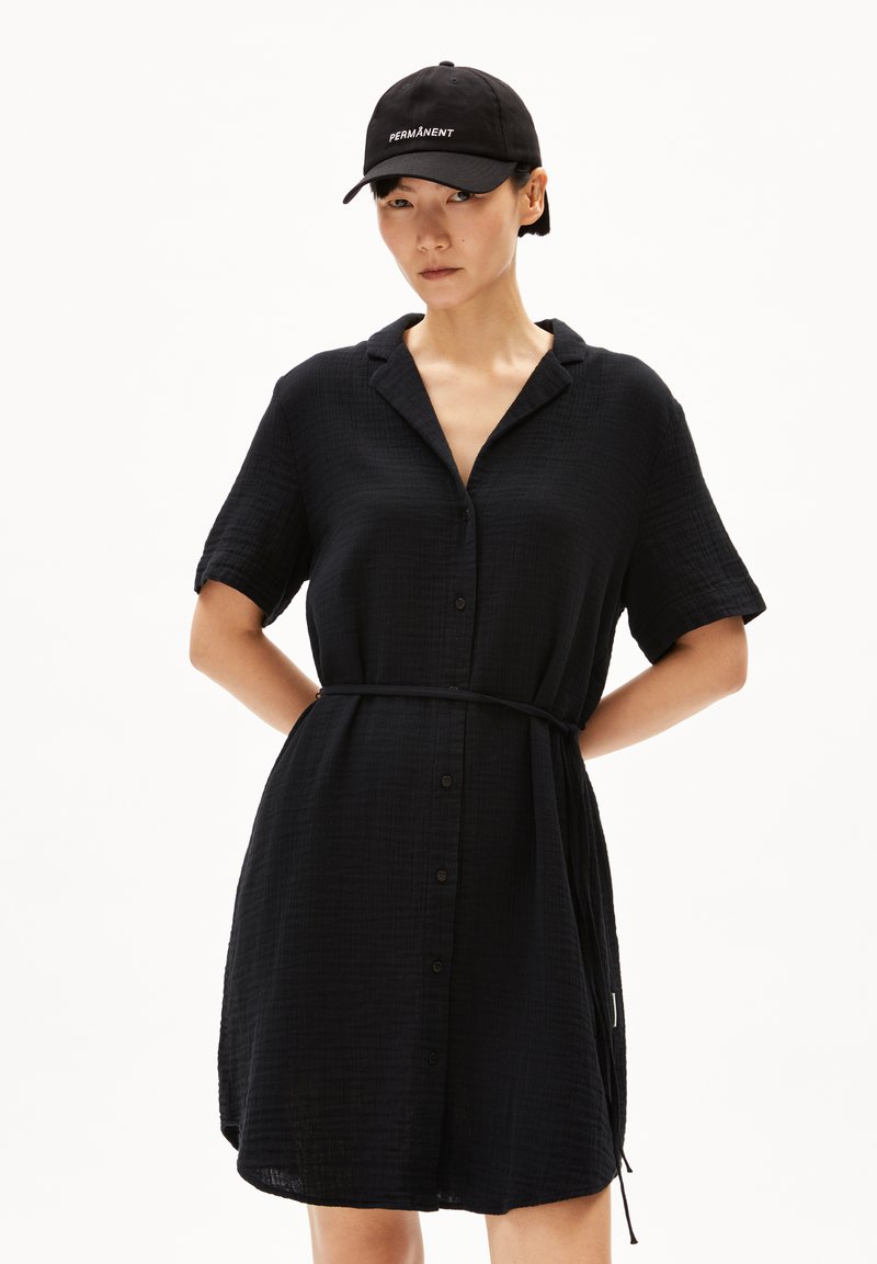 Schwarzes Hemdkleid mit kurzen Ärmeln, strukturiertem Stoff, Gürtel in der Taille und einem Kragen. Ergänzt durch eine schwarze Mütze mit weißem Text.
