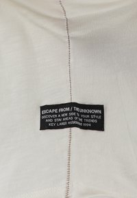 Tela blanca con un acabado texturizado, que presenta una etiqueta negra que dice "ESCAPAR DE / LO DESCONOCIDO" y detalles de costura a lo largo de la costura.
