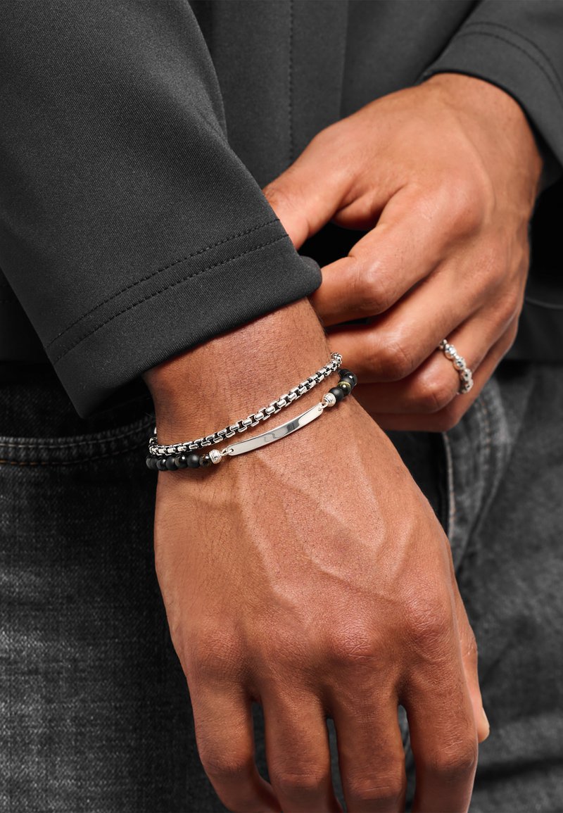 Silberarmband mit schwarzen Perlen, ausgestattet mit einem flachen Metallstück, einem Kettenglieder-Design und einer glatten Textur, getragen am Handgelenk.