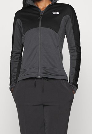 Kobieta w czarno-ciemnoszarej kurtce zapinanej na suwak z logo The North Face na piersi, z rękami w kieszeniach, na jednolitym tle.