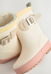Stivali alla caviglia in gomma color crema chiaro con una suola testurizzata rosa, dotati di una soffice fodera bianca, bordino giallo e un accessorio con fibbia sul lato.