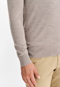 Jasno beżowy sweter z dzianiny z długimi rękawami, ściągaczami przy mankietach i u dołu. Miękka tekstura, dopasowany krój, w połączeniu z khaki spodniami widocznymi w talii.