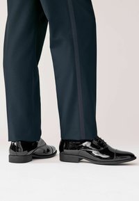 Schwarze Lacklederschuhe mit glänzender Oberfläche, runder Spitze und Schnürdesign, kombiniert mit dunklen Hosen.