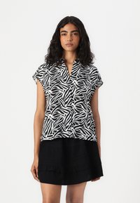 Chemise à manches courtes noire et blanche à motifs, en tissu léger, dotée d'un col et d'une ouverture à boutons. Portée avec une jupe noire à volants.