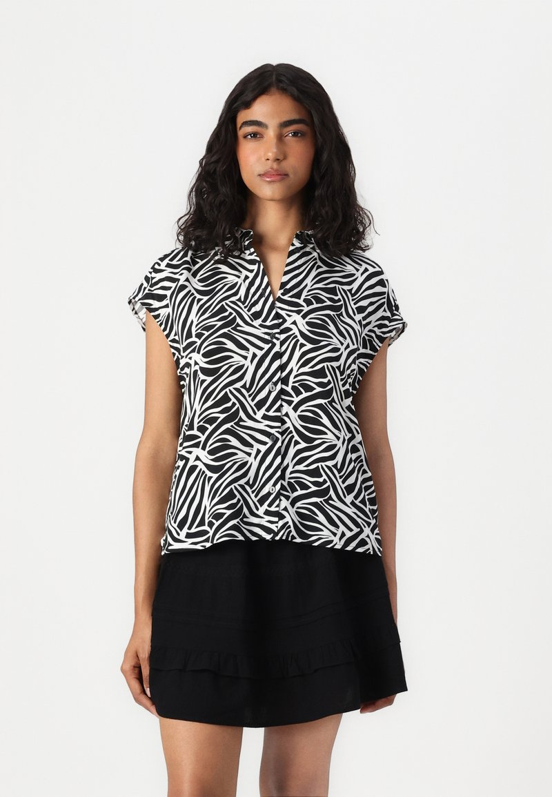 Chemise à manches courtes noire et blanche à motifs, en tissu léger, dotée d'un col et d'une ouverture à boutons. Portée avec une jupe noire à volants.
