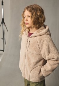 Jachetă fleece în bej deschis, cu închidere cu fermoar, glugă și buzunar frontal. Prezintă o suprafață texturată și o croială lejeră.