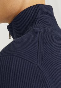 Col roulé en maille marine avec texture côtelée, coutures structurées et fermeture éclair au col pour plus de fonctionnalité.