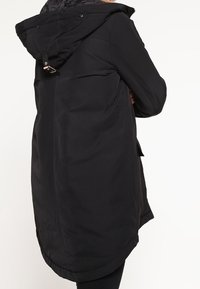 Černý svrchní oděv s kapucí, s hladkou texturou, dlouhým střihem a nastavitelnými kovovými doplňky vzadu. Jednoduchý design bez viditelných vzorů.