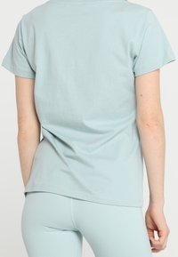 Osoba v světle modré tričko s krátkým rukávem a odpovídajícími legínami, zobrazená zezadu na jednobarevném pozadí.