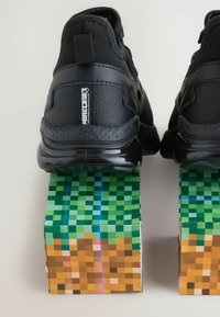 Baskets de sport noires avec un dessus texturé et des semelles brillantes, reposant sur des blocs pixelisés verts et bruns ressemblant à un paysage numérique.