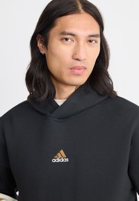 Črna kapucha s dolgimi rokavi, ki ima sprednji žep in vezeno zlato ter belo Adidasovo logo na prsih.