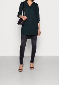 Mörkgrön långärmad tunika med någon slags krage, i kombination med svarta slim-fit byxor och svarta sandaler med öppen tå. Handväska med kedjehandtag.