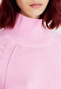 Pull rose en tricot avec un col montant côtelé, un tissu texturé et des manches raglan, mettant en valeur une finition douce et lisse.
