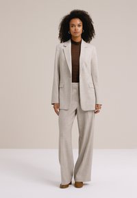 Kihara juustega naine, kellel on seljas beež ülikond laiade pükstega ja tumepruun mustriline särk, seisab ühevärvilise hele tausta ees.