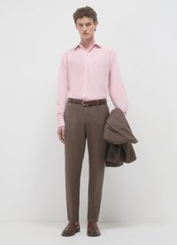 Hellrosa langärmeliges Hemd, braune Hose mit flacher Vorderseite, brauner Ledergürtel, mit einer passenden braunen Jacke über dem Arm gehängt. Braune Schuhe.