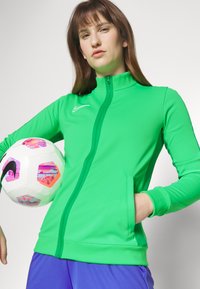 Grüne Zip-Jacke mit hohem Kragen, aus dehnbarem Stoff. Das Modell hält einen gemusterten Fußball und trägt lilafarbene Shorts.